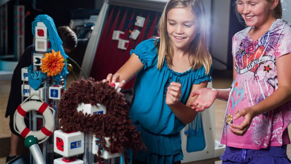 Two children interact with a small, colorful robot at a science lab table while other students look on.